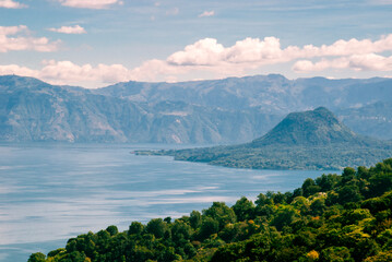 Scenic view of a lush forest and calm lake in Guatemala, with a green volcanic hill rising near the shoreline and vast mountains fading into the misty background under soft clouds.