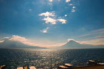 Scenic view of a lush forest and calm lake in Guatemala, with a green volcanic hill rising near the shoreline and vast mountains fading into the misty background under soft clouds.