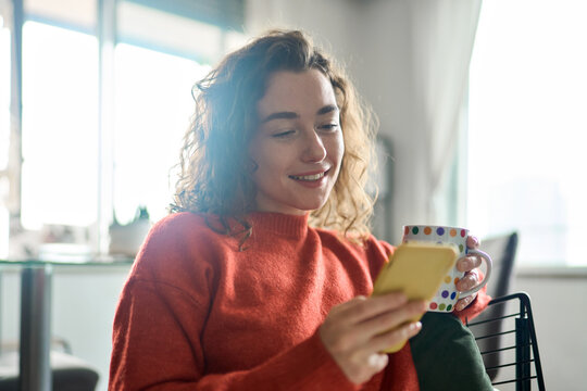 Smiling pretty lady holding smartphone enjoying mobile shopping, checking news on smartphone in cozy morning. Young happy woman using cell phone AI apps drinking coffee relaxing at home. - Powered by Adobe