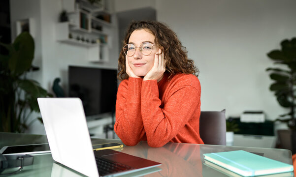 Pretty young woman student wearing glasses looking away thinking while working or learning online on laptop computer at home taking break, dreaming or contemplating, reflecting sitting at table.