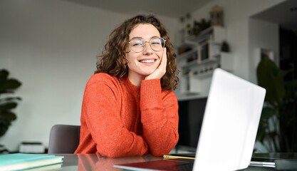 Portrait of happy young woman or student wearing glasses sitting at desk at home office with laptop computer looking at camera advertising online learning, remote work, business webinars.
