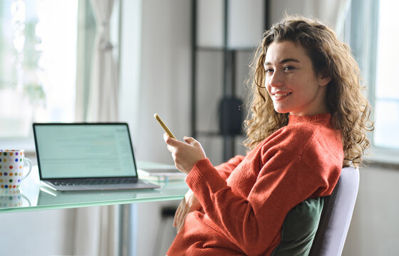 Happy calm lady feeling pleasure using phone at home. Young smiling pretty woman relaxing holding smartphone sitting in chair at table taking break after computer remote work or study. Candid photo