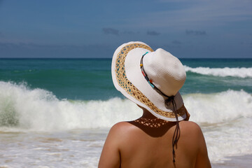 Tanned woman in sun hat standing on a beach on sea waves background. Summer travel and vacation