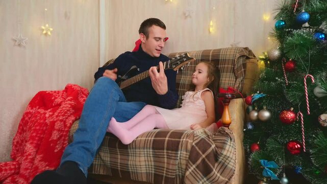 Father and little girl have fun together on Christmas holidays. Dad plays the guitar for his daughter.