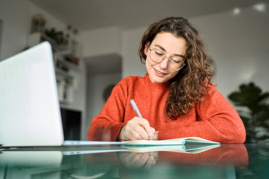 Pretty young woman student using laptop elearning or remote working at home office using laptop computer watching webinar, learning web course, studying online sitting at table, writing notes.