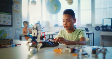 Primary School: African American Smart Boy Studying Robotic Arm Model During Science and Technology Lesson. Young Teacher with Tablet Computer Walking Around Modern Classroom. Creative STEM Education.