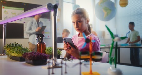 Talented Girl Sitting at Table and Using Smartphone, Conducting Biology Experiment on Plants with Tweezer and Magnifying Glass. Primary School Children During Lesson in Classroom. STEM Education.