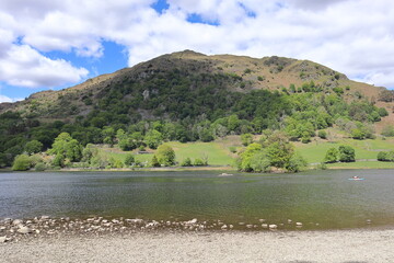 Shoreline of Rydal Water with rocks, calm water, greenery and surrounding hills under a partly cloudy sky. Lake District, England, UK.