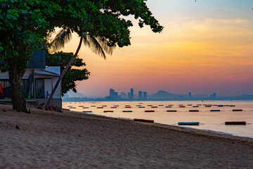 Fototapeta premium Tropical sunrise at Jomtien Beach, Pattaya, Thailand. Calm sea, warm colors in the sky, urban skyline in the background. Peaceful early morning coastal scene.