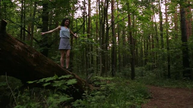 Happy joyful woman walking on fallen tree, jumping from log and spinning around on the path in the summer forest. Person coming on tree among the green trees in wood. Full length, fool height nature