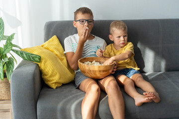 Two brothers sitting on gray sofa and watching movie while eating popcorn from wooden bowl at home...