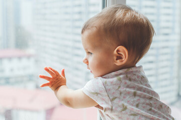 Curious baby infant in floral onesie looking out window and touching glass, concept of discovery, theme of childhood, early development, curiosity
