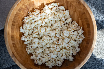 Freshly popped white popcorn in wooden bowl on soft carpet in natural sunlight at home. Concept of cozy movie night snack, homemade food, comfort lifestyle and casual indoor relaxation. 