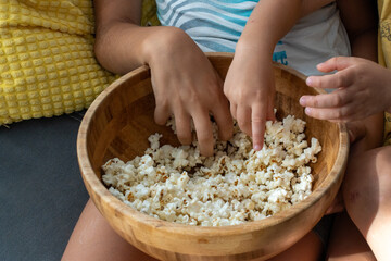 Close-up of children's hands reaching into wooden bowl of popcorn while sitting on couch at home. Concept of cozy family moment, snack time, childhood joy, and casual indoor relaxation together. 