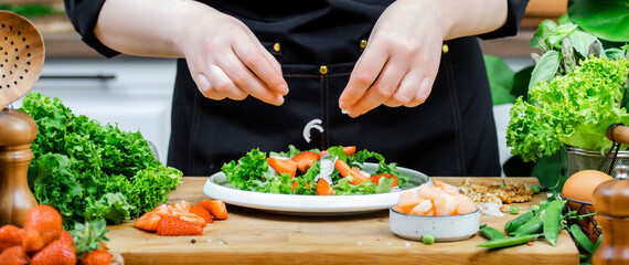 Hands of chef skillfully preparing colorful shrimp salad with fresh lettuce, strawberries, and other organic vegetables in bright kitchen. Healthy eating, cooking, food blogging