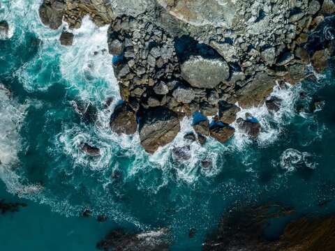 Aerial view of waves crashing against rocks in Big Sur, California, USA. The turquoise water contrasts with the rugged coastline, creating a dynamic scene.