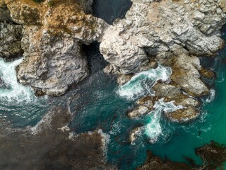 Aerial view of waves crashing against the rocky coastline of Big Sur, California, USA. The turquoise water contrasts with the rugged cliffs, showcasing the natural beauty.