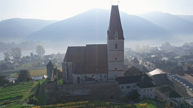Flight around of Weisenkirchen in der Wachau church at autumn morning with fog over Danube river. Wachau valley, Austria.