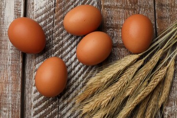 Raw eggs and spikes of wheat on wooden table, flat lay