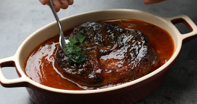 Woman adding pesto sauce to traditional ossobuco dish at grey table, closeup