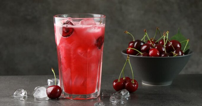 Pouring tasty cherry soda into glass with ice cubes on grey table, closeup