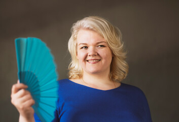 A chubby blonde woman fanning herself over grey background.