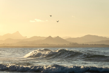 Waves roll in under a soft yellow haze with distant hills in Byron Bay.