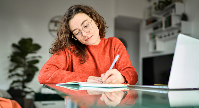 Busy young woman using laptop sitting at desk writing notes while watching webinar, studying online, learning web classes or having virtual call meeting remote working from home.
