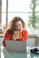 Naklejka na ściany i meble Vertical photo of young smiling pretty woman holding smartphone using cellphone modern technology, looking at mobile, checking cell phone apps, watching social media sitting at table at home.