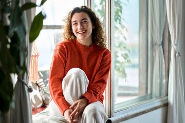 Happy pretty young positive charming woman sitting on windowsill relaxing at home looking at camera...