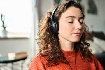 Young healthy mindful woman wearing headphones listening calming music audio podcast at home doing...