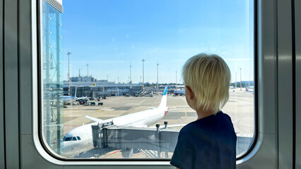 Horizontal portrait of Little blond boy looking out of the window in airport and curious watching...