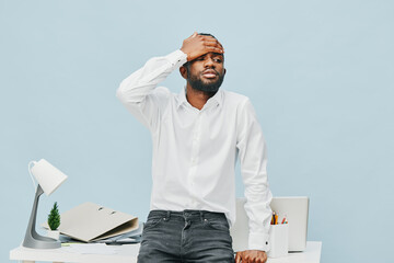 Frustrated young man with beard and afro hair, wearing white shirt, standing over a cluttered desk...