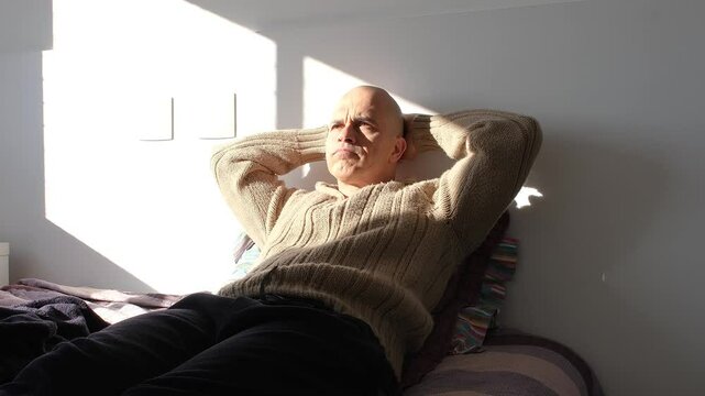 49 year old Brazilian man sitting on the bed deep in thought illuminated by sunlight.