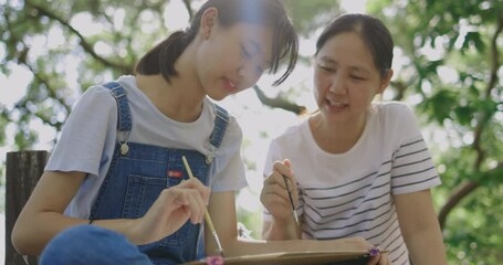 Slow motion Asian mother and daughter drawing a picture with paint and brush together in park backdrop.
