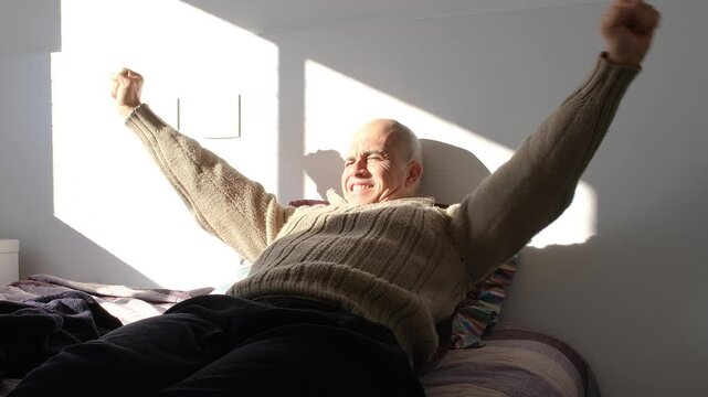 49 year old Brazilian man sitting on the bed waking up happy under the sunlight.