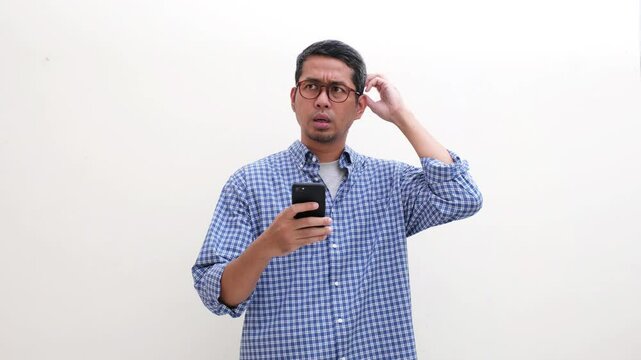 Adult Asian man showing confused expression and scratching his head after looking at his mobile phone