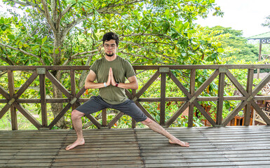Young man doing a yoga asana position in a forest wooden building with plants and green trees. Stretching exercise for good health, balance, positive mind, mental health and spiritual equilibrium