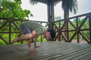 Young man doing a yoga asana position in a forest wooden building with plants and green trees. Stretching exercise for good health, balance, positive mind, mental health and spiritual equilibrium