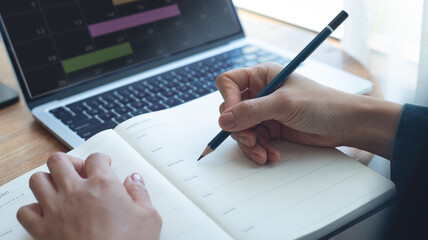 Businesswoman planning agenda and schedule, writing on calendar event planner notebook. Woman hand writing on plan reminder for work project and weekly event schedule on office table, close up