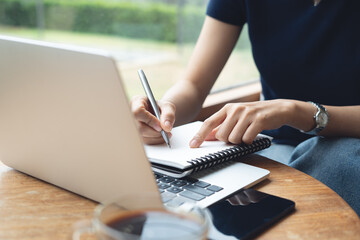 Business woman sitting at table in cafe, writing in notebook with laptop computer, mobile phone on...