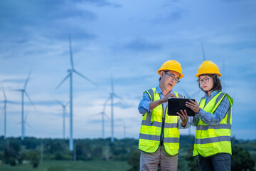 Two workers wearing safety vests and hard hats are looking at a tablet