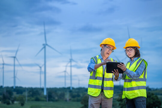 Two workers wearing safety vests and hard hats are looking at a tablet