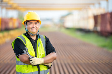 Confident Asian Engineer with Crossed Arms at Railway Construction Site Smiling Foreman in Hard Hat: Professional Worker at Train Depot Proud Industrial Technician Standing on Railway Tracks.