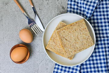 Top view of whole wheat sliced bread on plate with napkin and two chicken eggs, fork and knife. grey or gray cement as background. High angle, above, close up. blue, white.