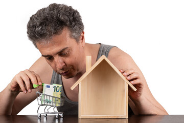 A poor middle-aged man in a sleeveless T-shirt, a miniature wooden house and a 100 euro note