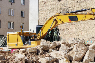 A large, yellow excavator located at a construction site among rubble and buildings.
