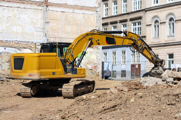 
A large, yellow excavator standing on a construction site with rubble, with old city buildings...