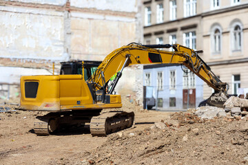 
A large, yellow excavator at a construction site, positioned near old building foundations or a demolished building and against the backdrop of city buildings.