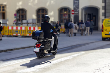 
A person riding a black scooter with a luggage box on the back, moving along a city street with...